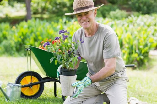 A middle-aged man in a light grey t-shirt, wearing a straw hat and gardening gloves, is kneeling on a well-maintained lawn in a residential garden. He is smiling while holding a potted plant with vibrant purple and red flowers, ready to plant or place it in the garden. Behind him, there is a bright green wheelbarrow with yellow wheels, partially filled with garden tools or materials, situated on grass. The background features a lush, leafy hedge and trees, indicating a landscaped outdoor space with natural light and clear weather, suitable for gardening activities. The scene suggests ongoing garden maintenance and planting, embodying professional outdoor care typical of local gardening services such as Lawn Mowing Yiewsley, serving the nearby area around Yiewsley and postcode UB7, with a focus on lawn and garden care.