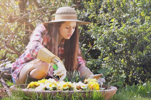 A young woman wearing a straw hat, a red and white checkered shirt, and gardening gloves is kneeling in a lush garden, tending to a flower bed filled with vibrant yellow, purple, and white flowers. Behind her, there are dense green bushes and trees, indicating a well-maintained outdoor space typical of a residential garden in Yiewsley. The garden features a grassy lawn and a raised flower bed edged with a stone border. The scene is illuminated by natural daylight, suggesting a clear, sunny day, and reflects a peaceful, tidy outdoor environment suitable for gardening and landscaping services offered by Lawn Mowing Yiewsley.